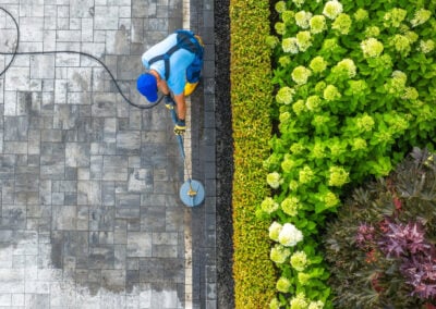 Worker pressure washing a tiled driveway next to a lush garden with green and colorful plants, viewed from above.